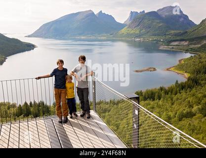 I bambini, in piedi sul bordo di una piattaforma panoramica lunga 44 metri, si affacciano sull'acqua del Bergsfjord, godendosi la vista sull'isola di Senja, nord nord nord Foto Stock