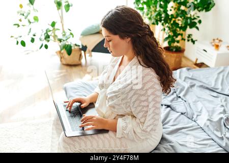 Una donna di grandi dimensioni si impegna con il proprio laptop seduto comodamente su un letto, circondato dal verde. Foto Stock