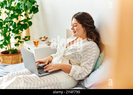 Una giovane e bella donna di grandi dimensioni si rilassa sul letto, usando felicemente il suo laptop a casa. Foto Stock