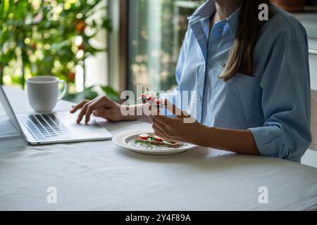 Donna tranquilla al tavolo la mattina che tiene un panino mentre fa colazione utilizzando un computer portatile. Foto Stock