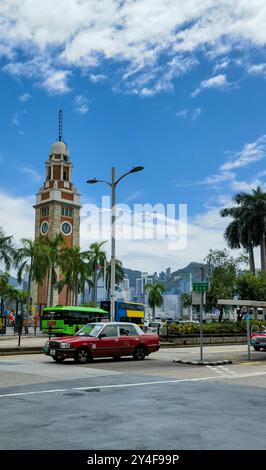 Hong Kong, Cina - 18 settembre 2024: Un taxi rosso è parcheggiato in un incrocio con una torre dell'orologio sullo sfondo. Le palme fiancheggiano la strada Foto Stock