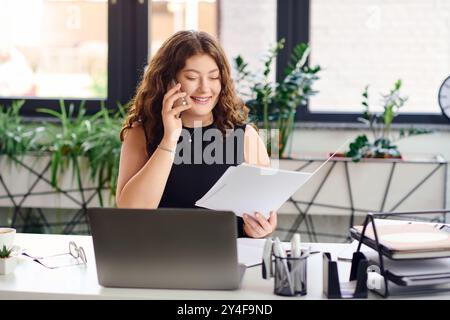 Una donna robusta e sicura di sé con lunghi capelli ricci sorride mentre esamina i documenti alla scrivania. Foto Stock