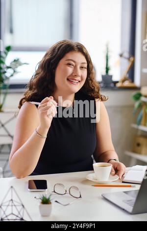 Una giovane donna sicura di sé con capelli ricci si impegna nel suo lavoro mentre si gusta una tazza di caffè alla sua scrivania. Foto Stock