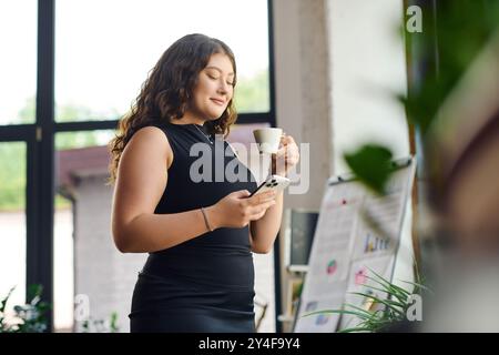 Una giovane professionista sicura di sé, con capelli ricci, assapora il caffè mentre interagisce con il suo smartphone in un ufficio vivace. Foto Stock