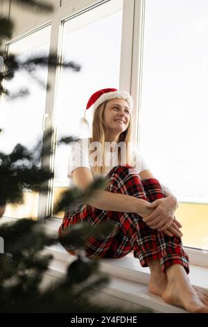 Donna di 35-40 anni siede sul davanzale vicino alla finestra e all'albero di Natale. C'è un cappello rosso di Babbo Natale sulla sua testa, è felice di sognare e sorridere. festi Foto Stock