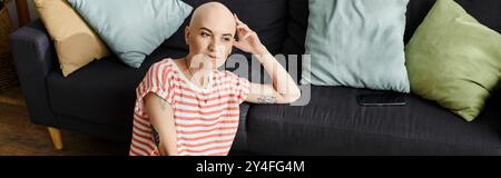 Una giovane donna con alopecia siede su un divano, guardando attentamente nel suo spazio colorato. Foto Stock