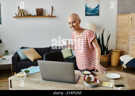 Una giovane donna allegra con alopecia sorseggia un caffè mentre si impegna con il suo laptop, trasudando calore. Foto Stock