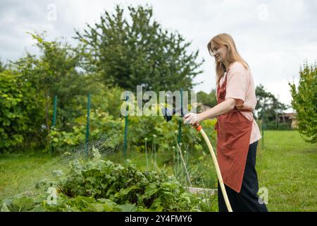 Donna felice che innaffia piante fresche che crescono nell'orto domestico. Giardiniere che si prende cura delle piante nel cortile di casa sua. Concetto di Foto Stock