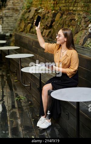 Una giovane donna con una gamba protesica sorride brillantemente mentre usa il suo telefono in un affascinante ambiente bar. Foto Stock