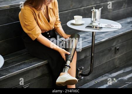 Una giovane donna siede comodamente in un bar, sorseggiando un caffè e regolando la gamba protesica, abbracciando la sua routine quotidiana. Foto Stock