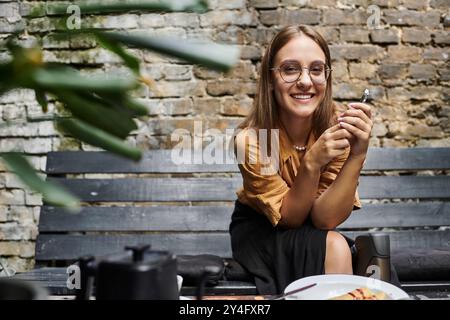 Una giovane donna siede in un bar, sorridendo mentre tiene in mano uno smartphone, mostrando la sua vita quotidiana e la sua fiducia. Foto Stock