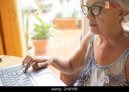 Una donna anziana si concentra mentre digita sul suo notebook, seduto accanto a una finestra in casa. Foto Stock