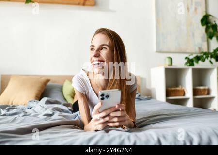 Una giovane donna sorride brillantemente mentre si rilassa sul letto, tiene il telefono e abbraccia un momento di felicità. Foto Stock