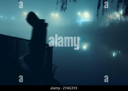 Un personaggio con cappuccio sfocato da solo. In piedi su una strada vicino a un fiume. In una misteriosa notte nebbiosa. In una città d'inverno. Foto Stock