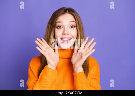 Foto ritratto di giovani affascinanti capelli biondi, simpatica ragazza sorridente con maglione arancione, palme sollevate sorprese isolate su sfondo di colore viola Foto Stock