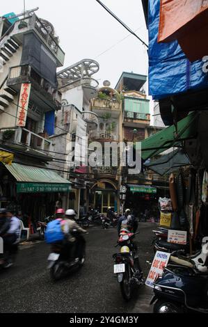 HANOI, Vietnam: Una vivace scena di strada di Hanoi mostra vecchi edifici che torreggiano su una strada stretta e affollata. Il vivace paesaggio urbano è pieno di moto, venditori ambulanti e pedoni che navigano nell'atmosfera vivace della città. Foto Stock