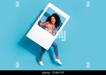 Foto a tutta lunghezza di una bella ragazza con cornice fotografica istantanea, vestito elegante e colorato, isolato su sfondo blu Foto Stock