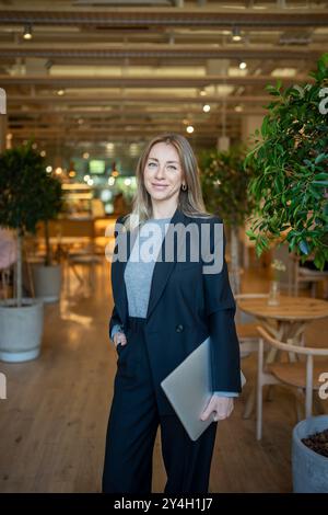 Una donna sicura di sé in costume da lavoro con un laptop in mano in piedi nel ristorante della hall guarda la fotocamera Foto Stock