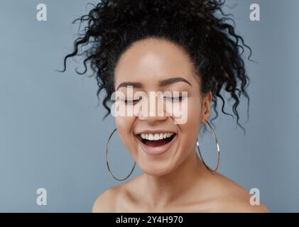 Ritratto di una donna che ride su sfondo grigio. Donna felice con capelli ricci e pelle pulita in posa in studio con gli occhi chiusi. Foto Stock