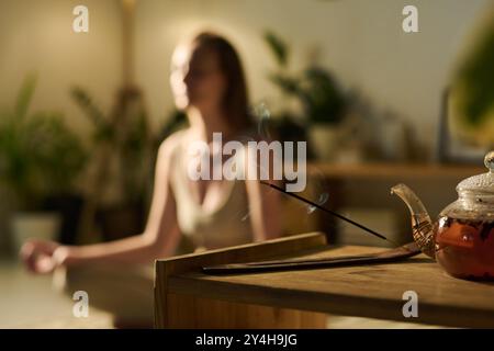 Parte di un tavolo in legno con tè alle erbe in teiera trasparente e supporto con bastoncino aromatico affumicato contro la giovane donna meditante Foto Stock