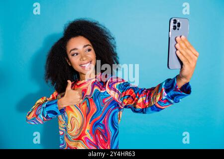 Foto ritratto di una bella ragazza scatta selfie foto con il pollice vestito elegante e colorato, con isolamento su sfondo blu Foto Stock