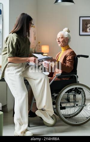 Donna anziana in sedia a rotelle con i capelli bianchi tirata indietro ricevendo documenti da una giovane donna in un ufficio domestico Foto Stock
