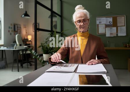 Ritratto di una donna d'affari anziana che esamina i documenti alla scrivania in un ufficio moderno con espressioni mirate, piante e lavagna bianca in background, mostrando d Foto Stock