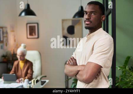 Ritratto di un giovane sicuro di sé in piedi con le braccia incrociate in un ufficio moderno con un collega anziano che lavora in background mantenendo la concentrazione mostrando seri Foto Stock