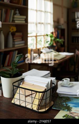 Luminoso ufficio domestico con ampia finestra che filtra in luce naturale, pieno di libri, piante, documenti cartacei e tazze da caffè, creando un ambiente di lavoro caldo e accogliente Foto Stock