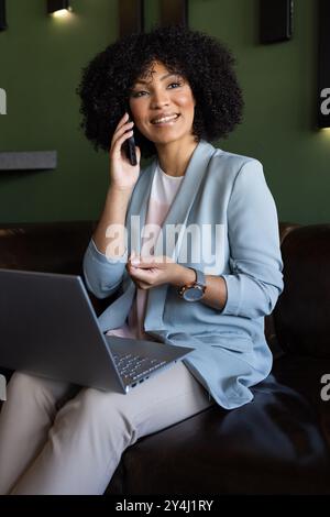 Parla con smartphone, donna che usa un laptop e sorride in ufficio Foto Stock