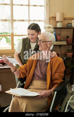Sorridente anziana donna caucasica in sedia a rotelle, assistita da un giovane assistente che tiene documenti con scaffali in legno e piante sullo sfondo Foto Stock