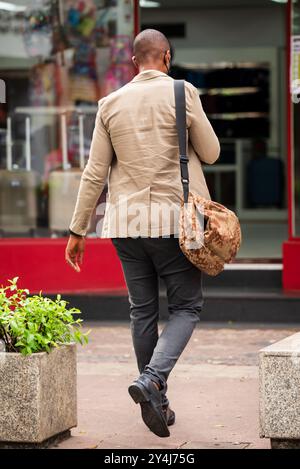 Vista posteriore dell'uomo nero brasiliano che indossa una giacca beige e una borsa mimetica che cammina verso un negozio di Savassi a Belo Horizonte. Foto Stock