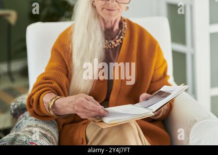 Donna anziana con lunghi capelli grigi che leggono un libro mentre è seduta comodamente sulla sedia a casa, mostrando un momento tranquillo e rilassato Foto Stock