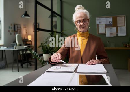 Ritratto di una donna d'affari anziana che esamina i documenti alla scrivania in un ufficio moderno con espressioni mirate, piante e lavagna bianca in background, mostrando d Foto Stock