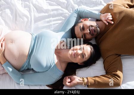A casa, coppia multirazziale, sdraiata sul letto, donna incinta e partner sorridenti e rilassanti insieme Foto Stock