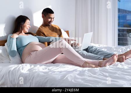 A casa, una coppia multirazziale incinta che si rilassa sul letto, utilizza un computer portatile e sorride insieme Foto Stock