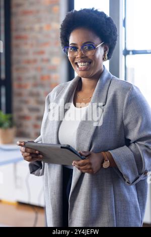 Porta un tablet, una donna d'affari sorridente in ufficio che indossa occhiali e blazer grigio Foto Stock