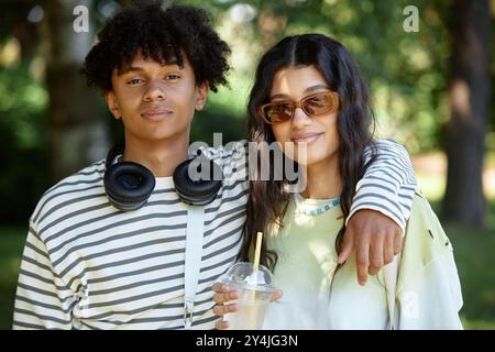 Ritratto di un adolescente afroamericano con le cuffie che posa accanto alla fidanzata del Medio Oriente che sorride per la fotocamera. Giovane coppia che si gode la giornata estiva nel parco Foto Stock