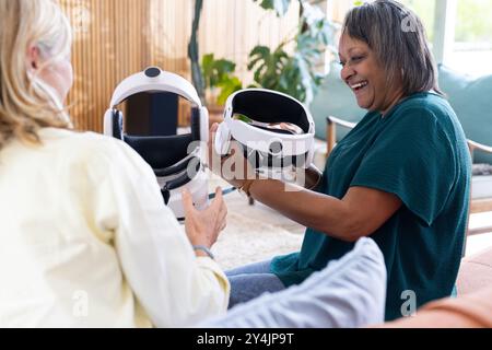 A casa, amiche di sesso femminile multirazziale che si godono gli occhiali per la realtà virtuale, sedute sul divano Foto Stock