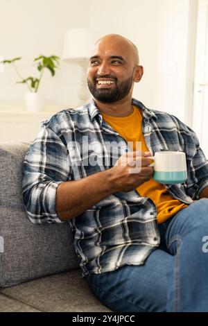 Uomo sorridente seduto sul divano tenendo in mano la tazza del caffè e godendosi un momento di relax Foto Stock