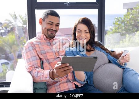 A casa, sorridere la coppia multirazziale incinta usando il tablet e tenendo la carta di credito, sul divano Foto Stock