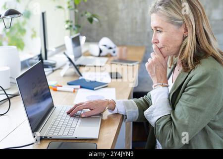 Lavora su un notebook, donna anziana che si concentra alla scrivania con forniture per ufficio Foto Stock