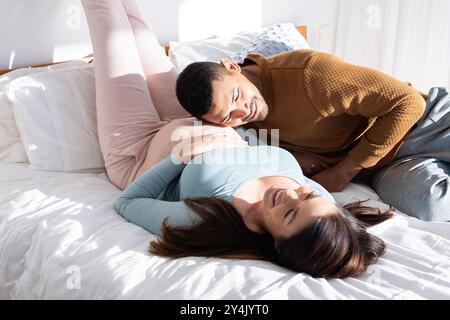 A casa, aspettandosi una coppia multirazziale che si rilassa sul letto, un uomo che tocca la pancia incinta della donna Foto Stock