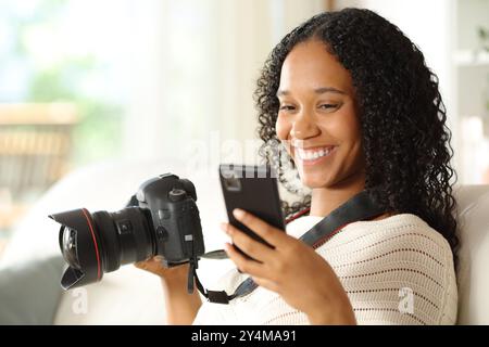 Felice fotografo nero che abbina il telefono alla fotocamera a casa Foto Stock