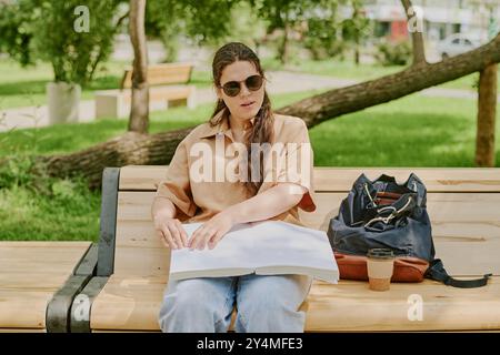 Giovane donna bruna con disabilità che indossa occhiali da sole e legge il romanzo in Braille mentre si gode una calda giornata nel parco locale Foto Stock