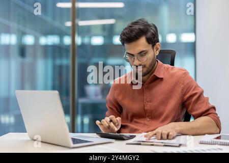 Giovane imprenditore maschile che lavora in ufficio utilizzando una calcolatrice e un computer portatile. Serio e incentrato sui calcoli finanziari, seduto al chiuso in un ambiente di lavoro moderno. Rappresenta il business e la produttività Foto Stock