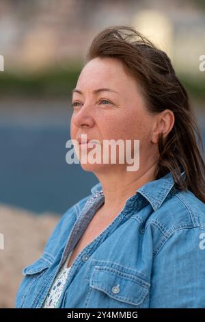 Ritratto di donna di mezza età. Alle donne piace la libertà. Seduti sulla riva del mare. Guarda con attenzione al mare. Riflessioni, tempo per se stessa Foto Stock