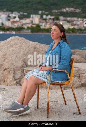 Ritratto di una donna felice di 50 anni seduta sulla sedia. Costa rocciosa. Goditi una vacanza al mare. Concetto di viaggio e vacanza Foto Stock