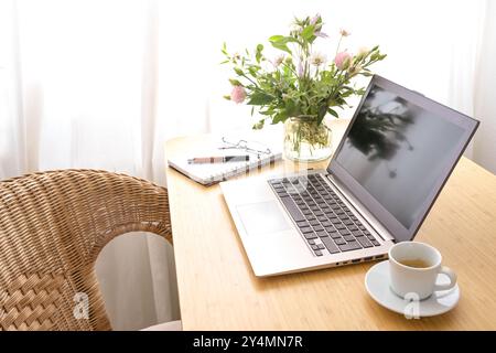 Computer portatile, caffè, bouquet di fiori e cancelleria su un tavolo di legno con poltrona in vimini, in un ufficio domestico caldo e accogliente, creativo Foto Stock