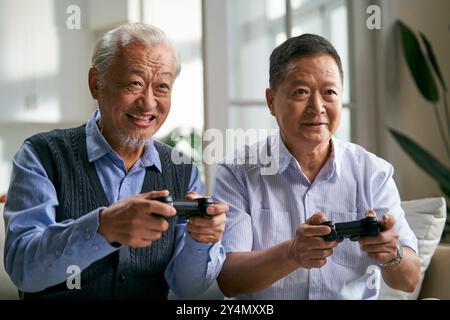 due uomini asiatici anziani felici seduti sul divano a casa a giocare a videogiochi insieme Foto Stock